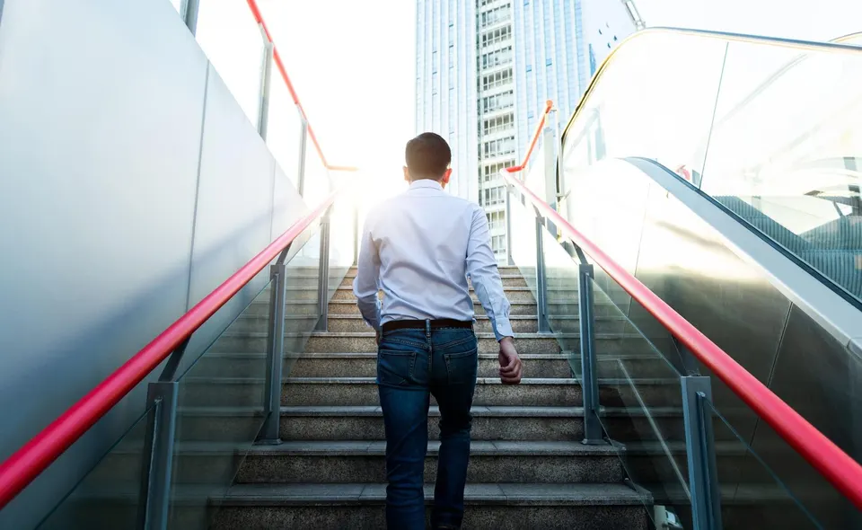Man in a white shirt and jeans walking up a stairway toward a bright cityscape, viewed from behind.